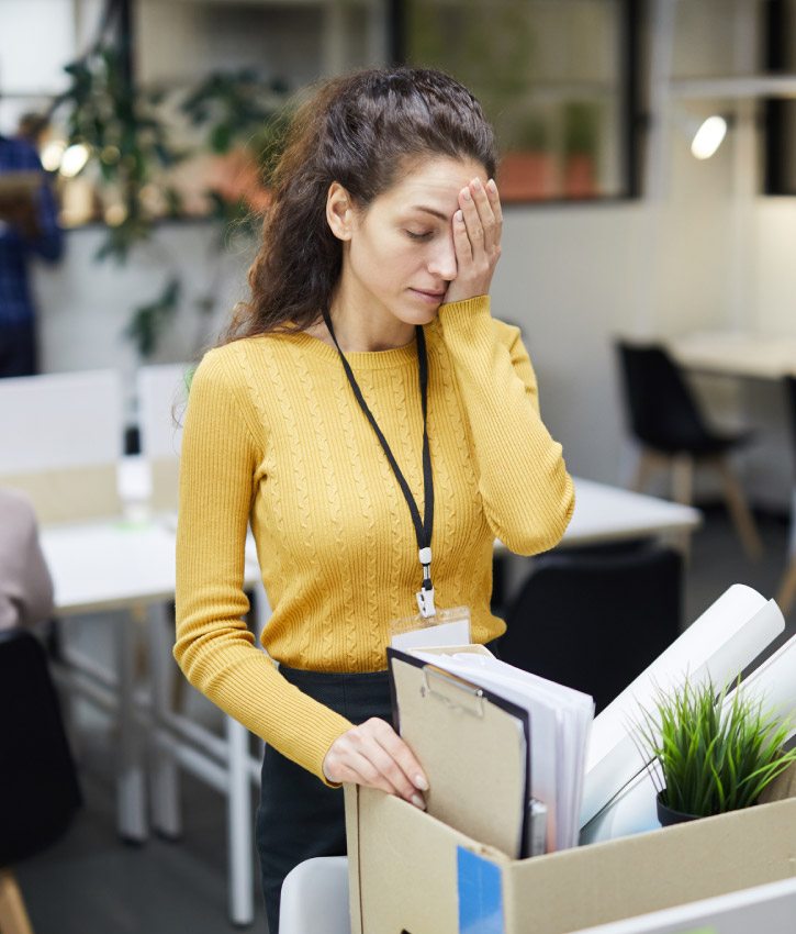 Frustrated young woman in yellow sweater standing at table and touching face with hand on her face while packing stuff in office after layoff