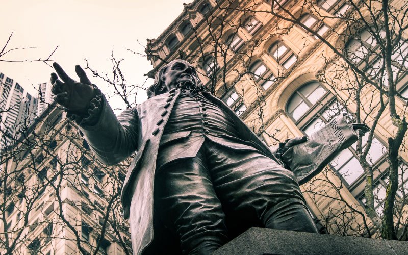 Upward view of a statue of Benjamin Franklin with tree branches and buildings on background