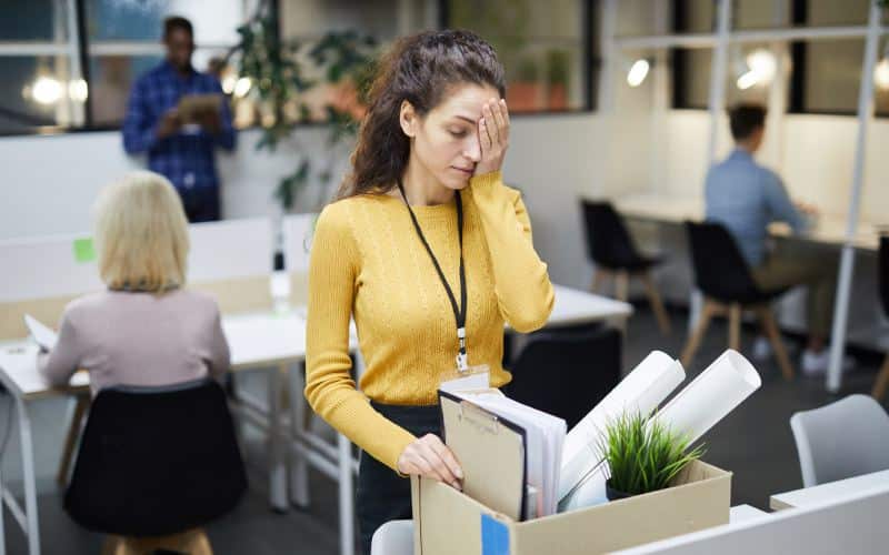 Frustrated young woman in yellow sweater standing at table and touching face with hand on her face while packing stuff in office after dismissal