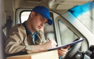 Young male delivery driver filling out form while sitting in van with package