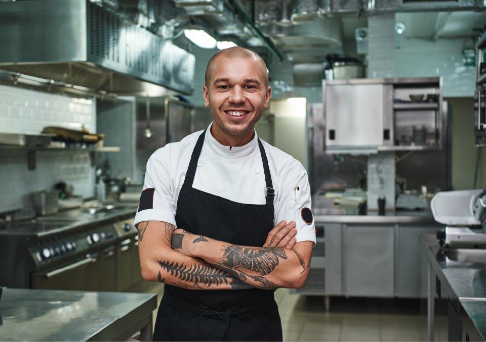 Cheerful young chef in apron keeping tattooed arms crossed and smiling while standing in a restaurant kitchen
